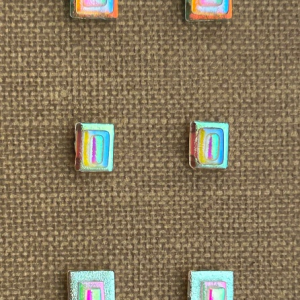 Three sets of square dichroic glass earrings with vibrant colors, arranged vertically on textured brown fabric for display