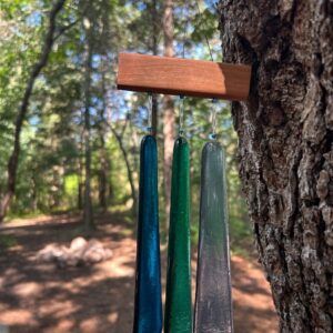Three fused glass wind chimes in blue, green, and clear hanging from wood mount on pine tree outdoors