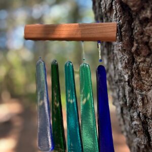 Side view of five fused glass wind chimes in blue, green, and clear mounted on pine tree with natural background