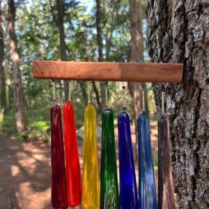 Seven multicolored fused glass wind chime blades in red, orange, yellow, green, blue, and purple mounted on wood in forest setting