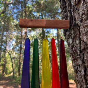 Five rainbow-colored fused glass wind chimes in purple, green, yellow, orange, and red attached to wooden hanger on pine tree