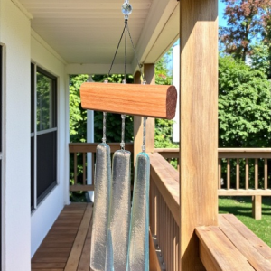 Glass wind chime with a wooden top hanging from the ceiling of a covered wooden porch on a sunny day